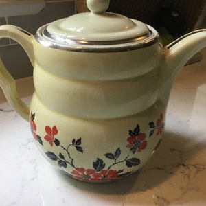 Puede incluir: Una tetera de cerámica amarilla vintage con un borde plateado y un diseño floral rojo y negro.