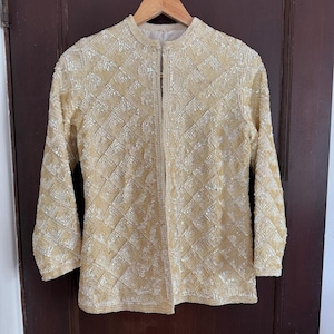 Pode incluir: Uma jaqueta vintage de manga comprida em cor dourada clara, coberta por pequenas lantejoulas brilhantes. A jaqueta tem gola redonda e abertura vertical na frente. O padrão de lantejoulas cria um design em forma de diamante na peça.
