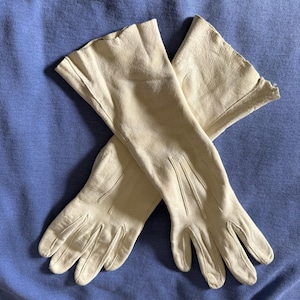 May include: A pair of cream-colored leather gloves with a smooth texture and a matte finish. The gloves feature a mid-length design that extends past the wrist, with three vertical stitched lines on the back of each hand and a scalloped edge.