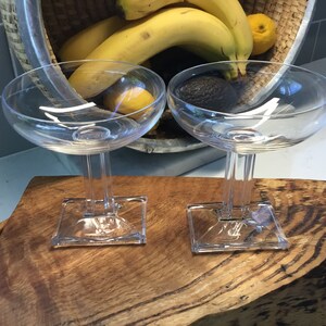 May include: Two clear glass champagne coupes with square bases sit on a wooden surface. The glasses are empty and appear to be made of crystal.