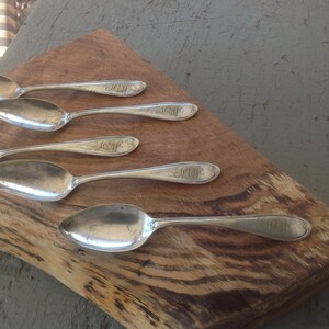 May include: Five silver spoons with ornate handles, laid out on a wooden surface. Each spoon has an engraved monogram.