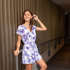 May include: A woman wearing a white and purple floral print short dress with a V-neckline and short sleeves. She is standing in a hallway with a metal railing.
