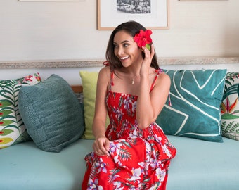 May include: A woman in a red floral maxi dress sits on a light blue couch with green and white patterned pillows. She is wearing a flower behind her ear and smiling.