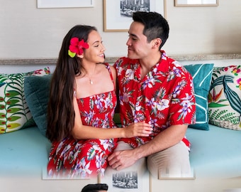 May include: A couple wearing matching red floral print shirts and dresses pose on a blue couch. The woman is wearing a flower in her hair.