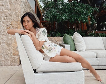 May include: A woman wearing a white floral shirt and white shorts is sitting on a white couch with a green pillow. The couch is on a patio with a stone wall and green plants in the background.