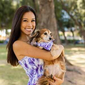 以下が含まれることがあります： 白と紫の花柄のドレスを着た女性が、茶色と白の小さな犬を抱っこしています。犬は花柄の紫色のバンダナをしています。