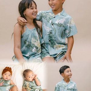 Pu&ograve; includere: Una famiglia di quattro persone vestite con abiti coordinati a stampa tropicale. I due figli maggiori sorridono e si guardano. I due figli pi&ugrave; piccoli sono seduti su un letto bianco. Gli abiti sono di colore azzurro chiaro con foglie verdi e fiori bianchi.