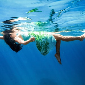 May include: A woman floats in a pool of clear blue water, wearing a green and white patterned dress. Her hair is dark and flowing, and she appears to be relaxed and enjoying the water.