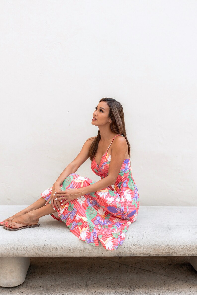May include: A woman wearing a pink and white floral dress sits on a concrete bench. She is looking up and to the right.