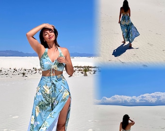 May include: A woman wearing a blue and white floral print two-piece swimsuit with a halter top and a high-waisted skirt with a thigh-high slit. She is standing in a white sand desert with a blue sky and white clouds in the background.