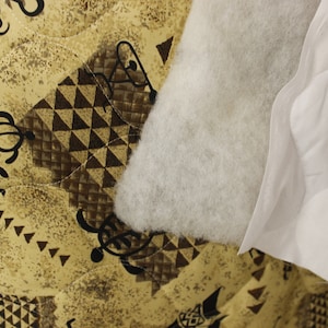 May include: Close-up of a white quilted fabric with a brown and tan patterned fabric underneath. The white fabric has a fluffy texture and the patterned fabric has a geometric design.