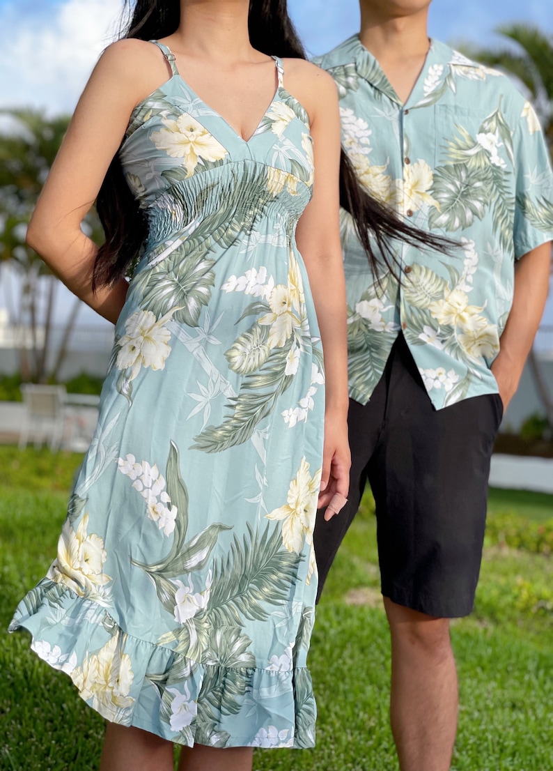May include: A couple wearing matching tropical print clothing. The woman is wearing a light blue sleeveless dress with a floral pattern. The man is wearing a light blue short-sleeved shirt with a floral pattern and black shorts.