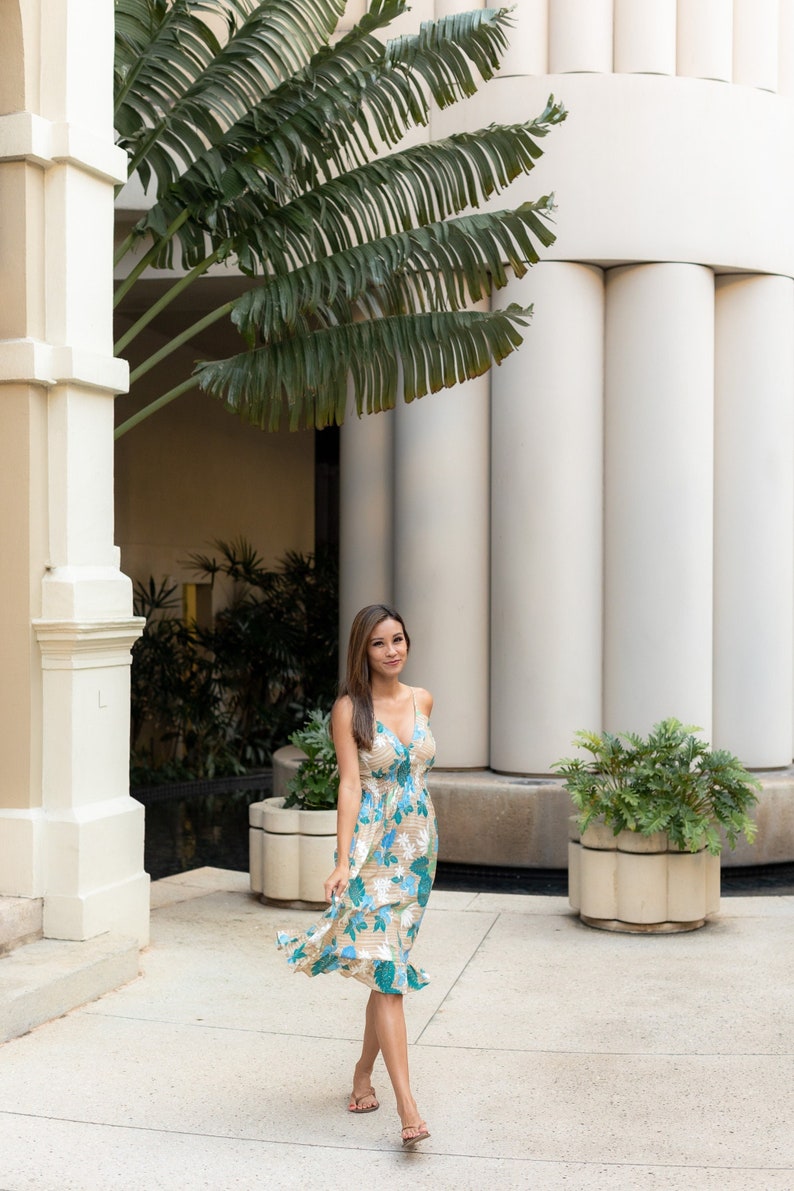 May include: A woman in a floral dress walks in a courtyard with white columns and palm trees.