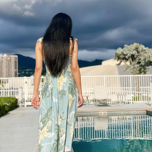 May include: A woman with long black hair walks towards a swimming pool wearing a light blue floral maxi dress.