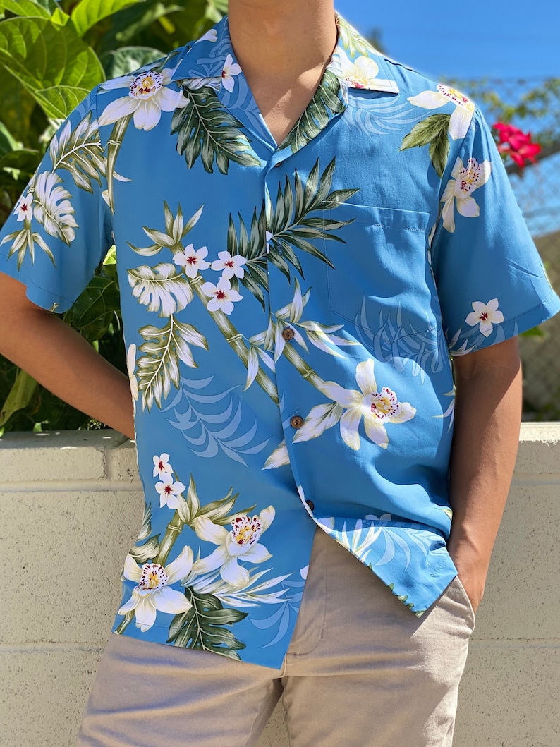 K&ouml;nnte beinhalten: Ein hellblaues Kurzarmhemd mit einem wei&szlig;en Blumenmuster. Das Hemd hat einen Knopfkragenshirt und eine Tasche auf der linken Seite.