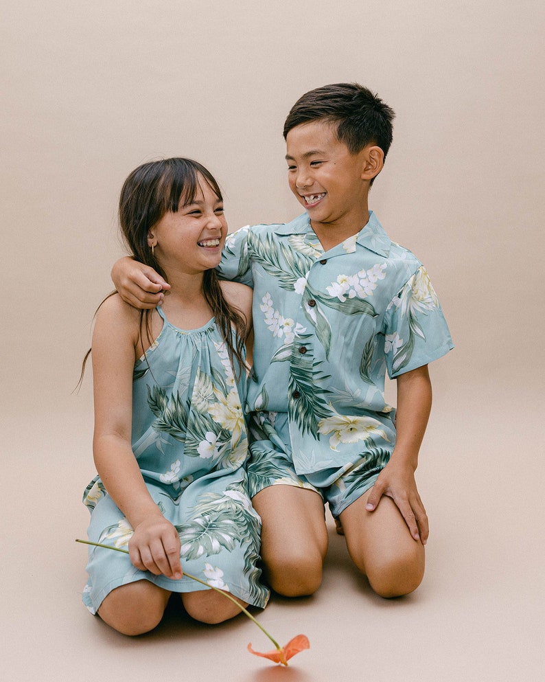 May include: Two children, a boy and a girl, wearing matching blue and white floral print clothing. They are sitting on the floor and smiling at each other. The girl is holding a single orange flower.