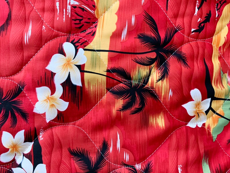 May include: Red fabric with a tropical print featuring palm trees, white flowers, and a sunset scene. The fabric has a quilted texture.