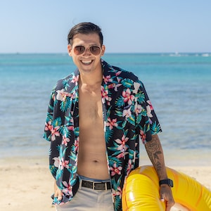 May include: A man wearing a black and pink floral shirt and white shorts is standing on a beach holding a yellow inflatable ring. He is smiling and wearing sunglasses.