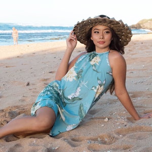 May include: A woman wearing a blue floral halter dress and a wide-brimmed straw hat sits on a sandy beach.