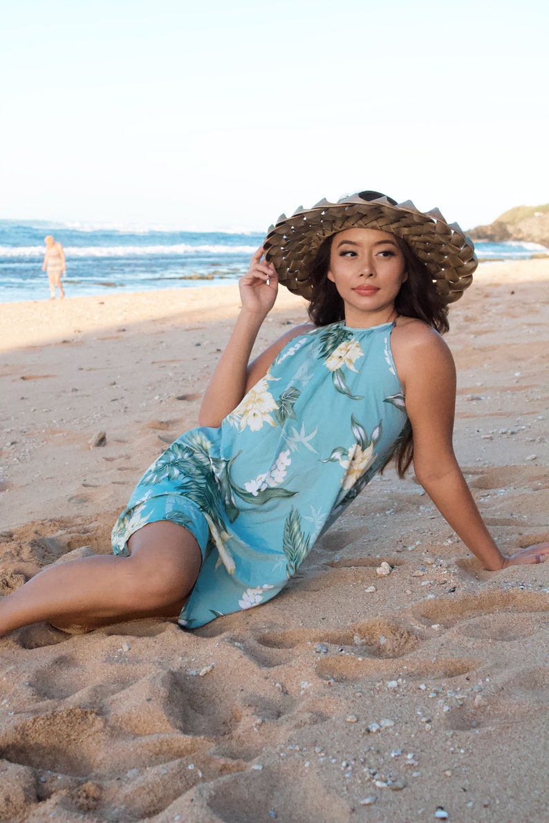 May include: A woman wearing a blue floral halter dress and a wide-brimmed straw hat sits on a sandy beach.