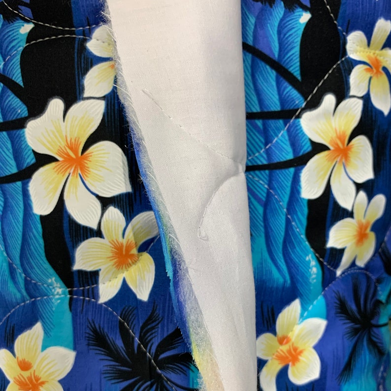 May include: Close-up of a blue and white fabric with a tropical print featuring palm trees and white flowers. The fabric is quilted and has a white backing.