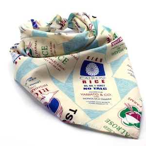 May include: A bandana with a cream background and blue, red, and green accents. The bandana features a repeated pattern of rice bags with the text "Calrose Rice US. No. 1-Fancy No Talc Packed for Yamato & Co. Ltd. Honolulu Hawaii Coated with Edible Food Products" and "Kiku Rice" with a Japanese flower symbol.