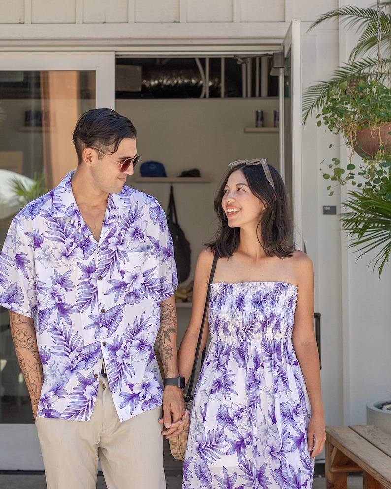 以下が含まれることがあります： 白と紫の花柄のシャツを着たカップル。男性は襟と半袖の白いシャツを着ています。女性はストラップレスのドレスを着ています。彼らは白い建物の入り口の前に立っています。