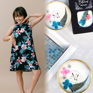 May include: A young person wearing a black and floral print dress. The image also shows two wax sachets with dried flowers and greenery inside. One sachet has the text "Ninth Isle" and the other has the text "Botanical Wax Sachet".