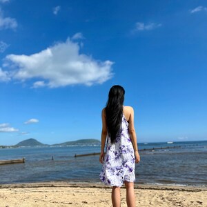 以下が含まれることがあります： 白い砂浜に立って、海を見つめる女性。彼女は白地に紫の花柄のワンピースを着ています。空は青く、白い雲が浮かんでいます。