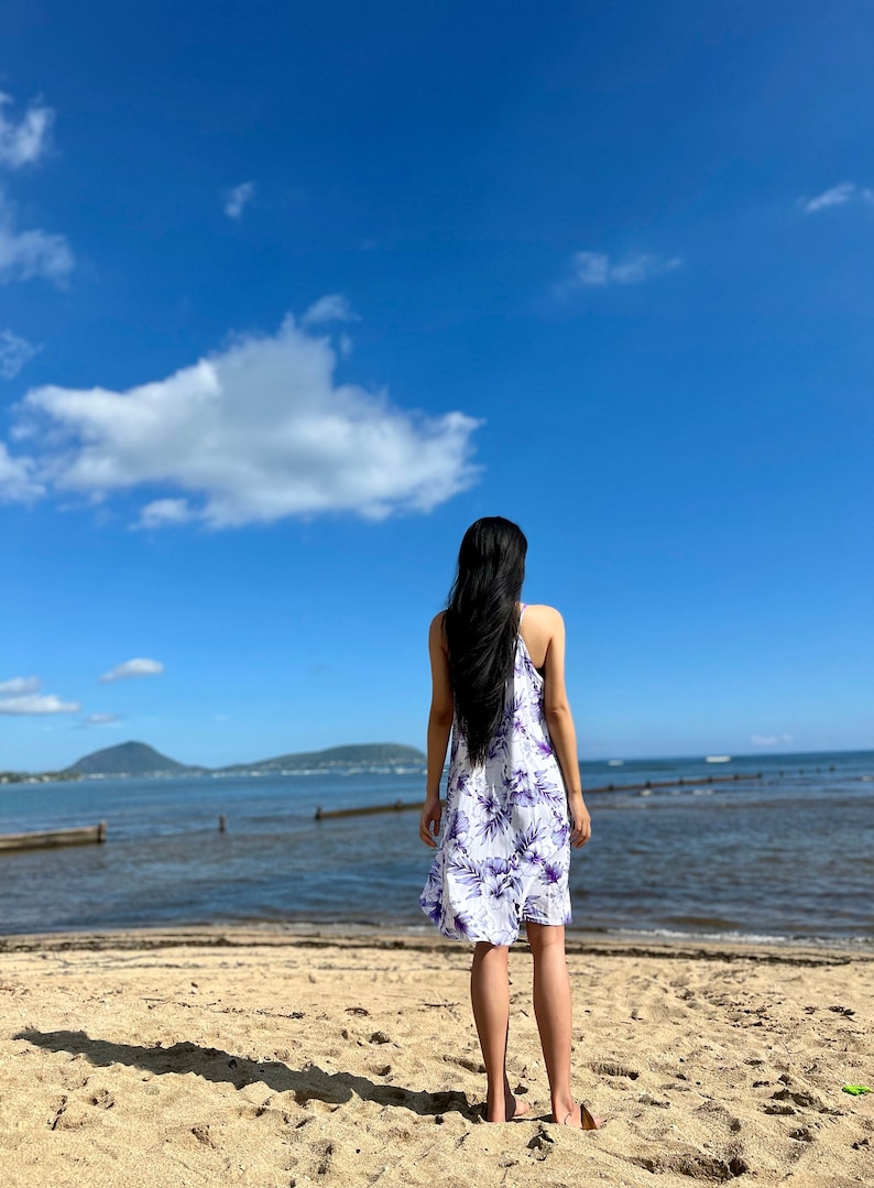 以下が含まれることがあります： 白い砂浜に立って、海を見つめる女性。彼女は白地に紫の花柄のワンピースを着ています。空は青く、白い雲が浮かんでいます。