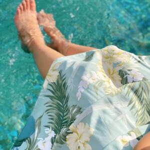 May include: A person's legs are in a pool of water, wearing a light blue skirt with a tropical floral print. The skirt has green leaves, white flowers, and yellow flowers.