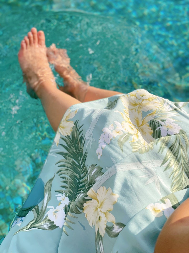 May include: A person's legs are in a pool of water, wearing a light blue skirt with a tropical floral print. The skirt has green leaves, white flowers, and yellow flowers.