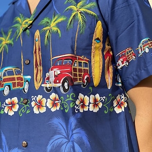 K&ouml;nnte beinhalten: Ein blaues, kurz&auml;rmeliges Hemd mit Kn&ouml;pfen und einem tropischen Druck mit Palmen, Surfbrettern und Oldtimern. Das Hemd hat einen wei&szlig;en Hibiskus-Bl&uuml;tenrand.