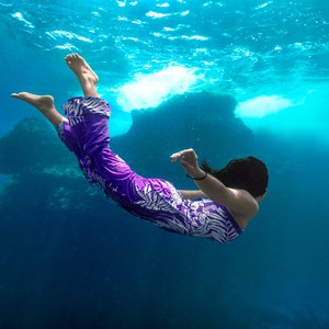May include: A person in a purple and white floral patterned dress swims underwater. The person is floating with their arms outstretched and legs extended. The water is clear and blue.