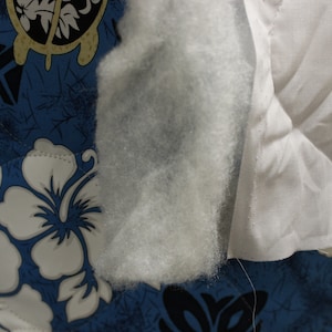 May include: Close-up of white cotton fabric with a textured surface. The fabric is folded and has a seam along the edge. The fabric is on top of a blue and white patterned fabric.