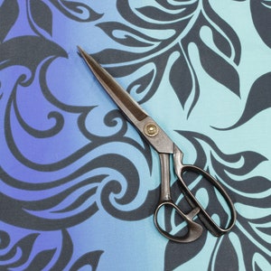 May include: A pair of antique silver scissors with a black handle, lying on a blue and black patterned fabric.
