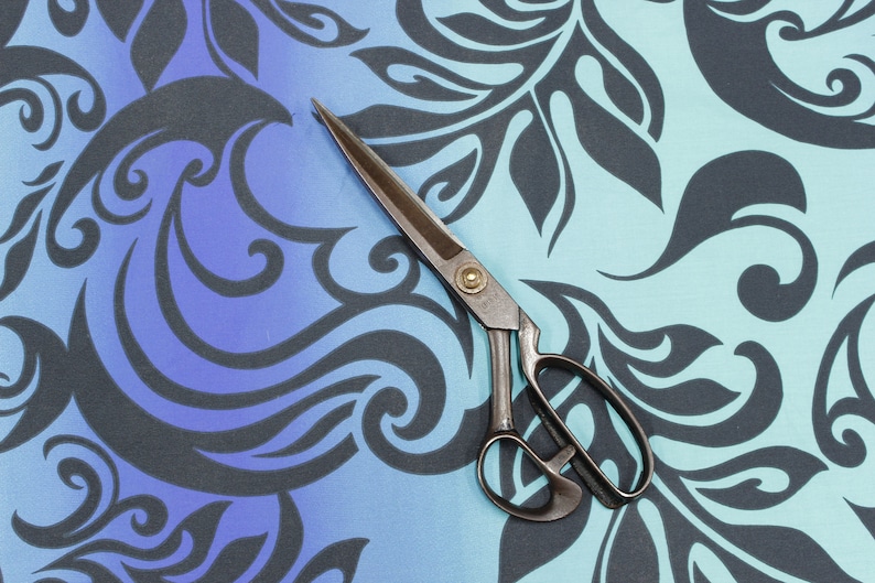 May include: A pair of antique silver scissors with a black handle, lying on a blue and black patterned fabric.