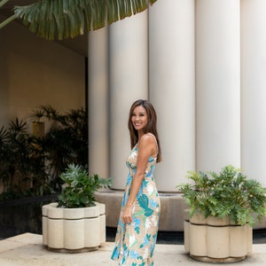 May include: A woman wearing a floral print midi dress with a light brown background and blue and green floral accents. She is standing in front of a white building with columns and green plants.