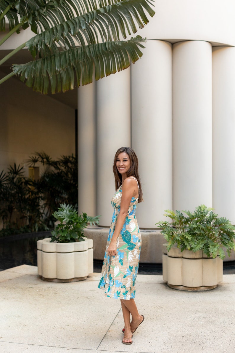 May include: A woman wearing a floral print midi dress with a light brown background and blue and green floral accents. She is standing in front of a white building with columns and green plants.