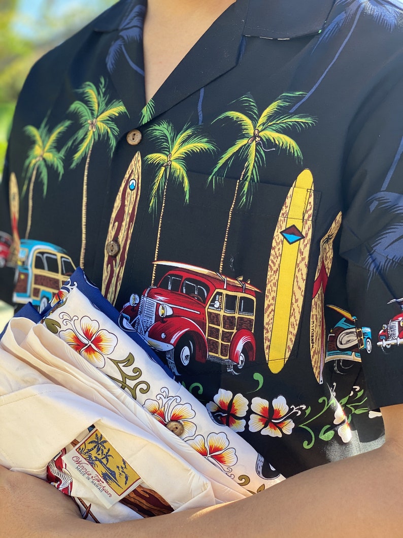 K&ouml;nnte beinhalten: Ein schwarz-wei&szlig;es Hawaii-Hemd mit Palmen, Surfbrettern und einem alten Holz-Kombi. Das Hemd ist gefaltet und wird von einer Person gehalten. Das Hemd hat ein Etikett mit der Aufschrift "Charlie Trotter, Made in Hawaii".
