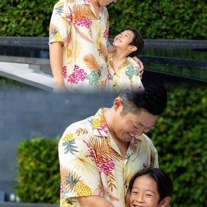 May include: A father and son wearing matching yellow, green, and red patterned shirts with tropical leaf designs. The father is smiling down at his son, who is smiling up at him. They are standing in front of a pool.