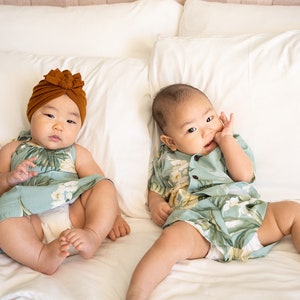 May include: Two babies wearing matching tropical print outfits lay on a white bed. One baby is wearing a brown turban and the other is wearing a short-sleeved shirt.