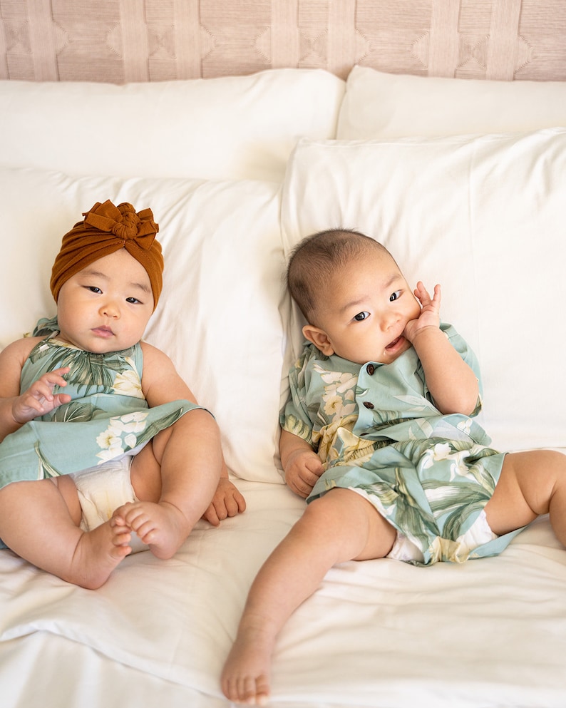 May include: Two babies wearing matching tropical print outfits lay on a white bed. One baby is wearing a brown turban and the other is wearing a short-sleeved shirt.