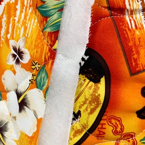 May include: Close-up of a white fabric seam between two pieces of orange and yellow fabric with a tropical print. The fabric has a quilted texture and features a hibiscus flower and palm leaves. The text "HAWAII OAHU" is visible on the right side of the image.