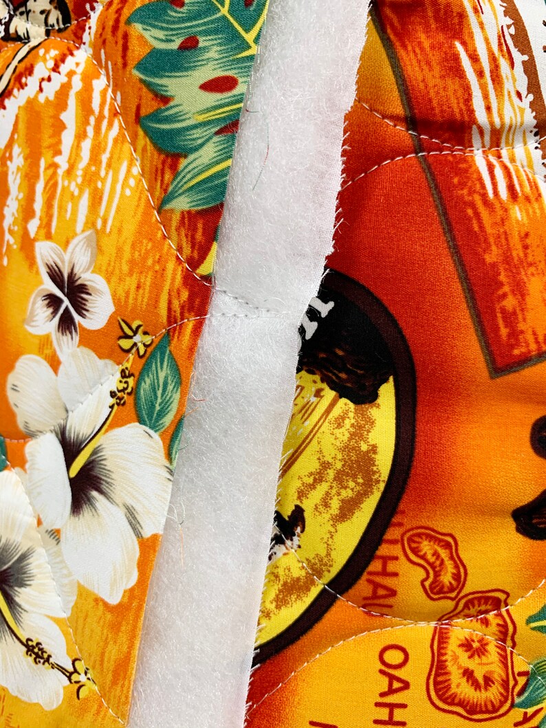 May include: Close-up of a white fabric seam between two pieces of orange and yellow fabric with a tropical print. The fabric has a quilted texture and features a hibiscus flower and palm leaves. The text "HAWAII OAHU" is visible on the right side of the image.