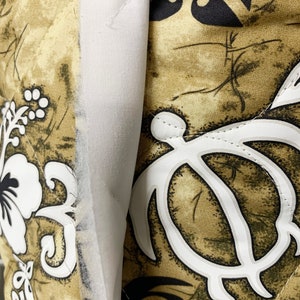 Op de afbeelding: Een close-up van een bruine en witte stof met een gestikte textuur. De stof heeft een herhalend patroon van gestileerde schildpadschalen, bloemen en spiralen. De stof is begrensd door een witte strook stof.
