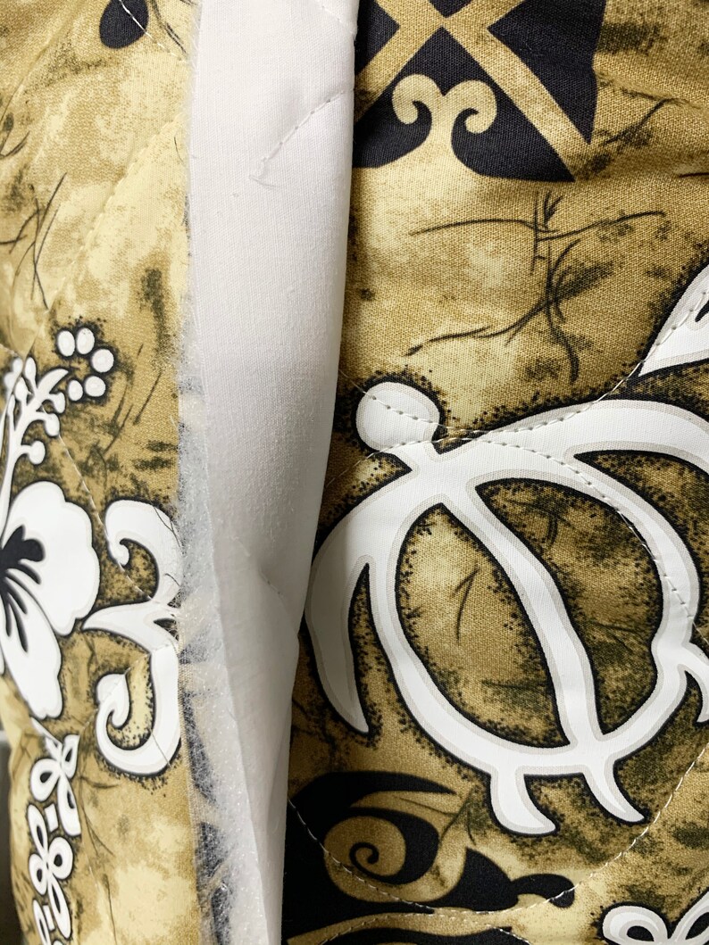 Op de afbeelding: Een close-up van een bruine en witte stof met een gestikte textuur. De stof heeft een herhalend patroon van gestileerde schildpadschalen, bloemen en spiralen. De stof is begrensd door een witte strook stof.