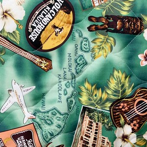 May include: A green quilted fabric with a Hawaiian theme. The fabric features images of a ukulele, an aeroplane, a map of the Hawaiian islands, and tropical flowers. The text "Volcano House 2 Kilauea" is visible on the fabric.