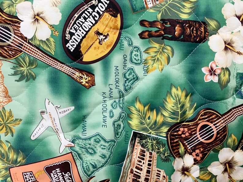 May include: A green quilted fabric with a Hawaiian theme. The fabric features images of a ukulele, an aeroplane, a map of the Hawaiian islands, and tropical flowers. The text "Volcano House 2 Kilauea" is visible on the fabric.