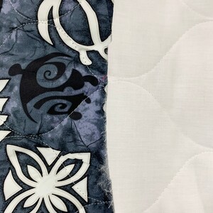 Op de afbeelding: Een close-up van een grijze en witte stof met een gestikt patroon. De stof heeft een herhalend ontwerp van gestileerde bloemen en schildpadden. De stof is genaaid aan een witte stof met een gestikt patroon.